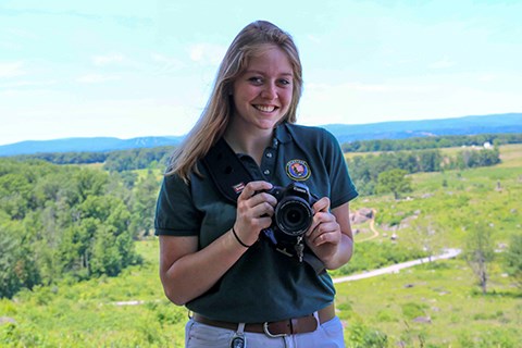 A female intern in a green shirt holds a camera with Devil's Den in the background.