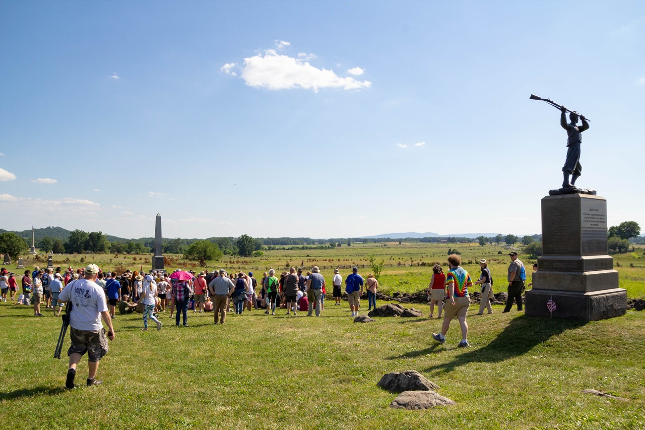 A large crowd of visitors gather near a low stone wall in front a field with wooden fences in the background and three granite and bronze monuments in the foreground.