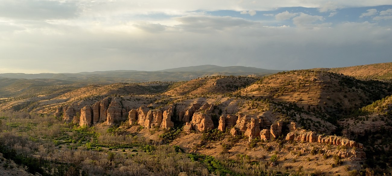 An overlook of the Gila Wilderness featuring large spires of rock with green vegetation in the foreground with rolling hills stretching out into the background.