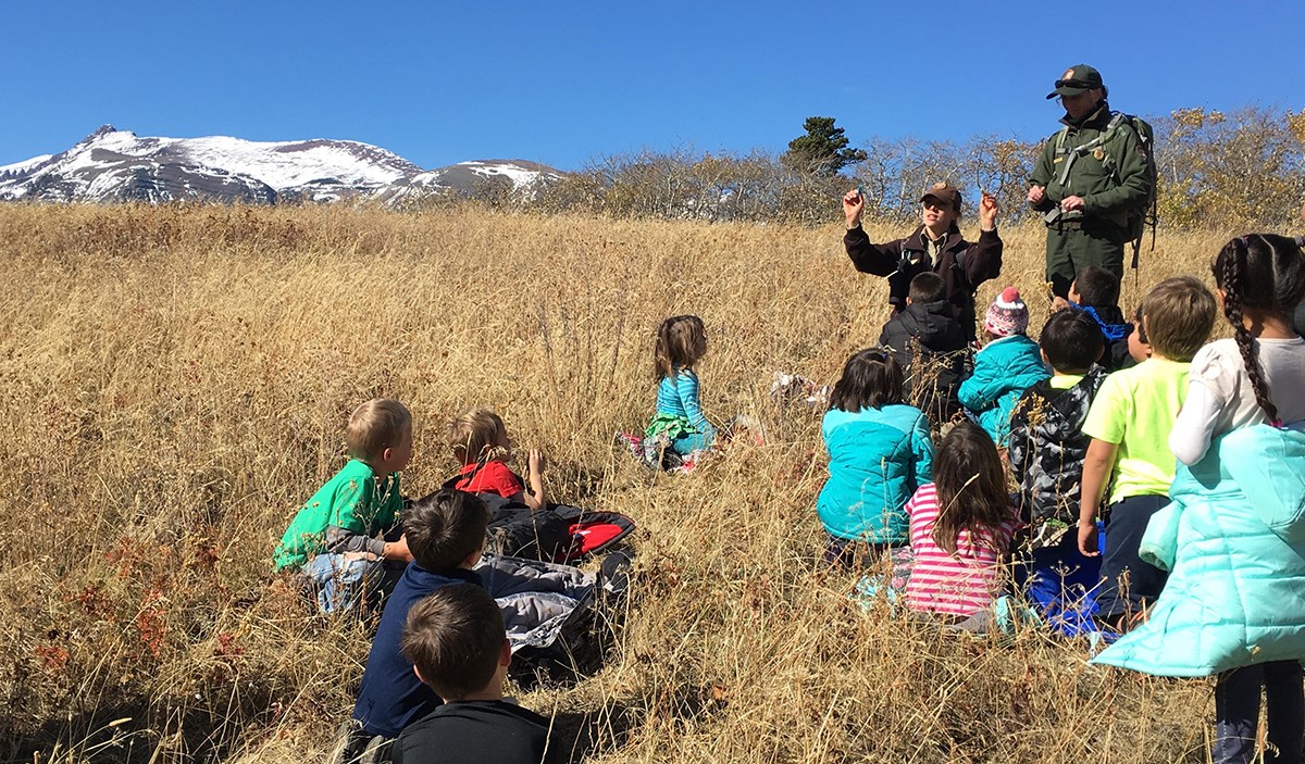 Students sit in tall prairie grass while listening to kneeling ranger talking and holding up her hands with another ranger standing behind