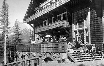 historic image of people in period dress on porch of large wooden building with mountains in background