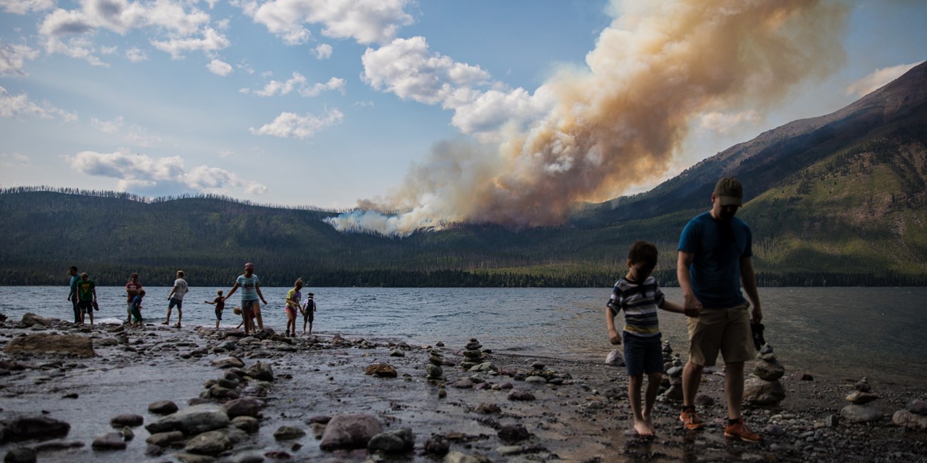 Families walk on a cobble beach and a wildland fire burns in the background across a lake.