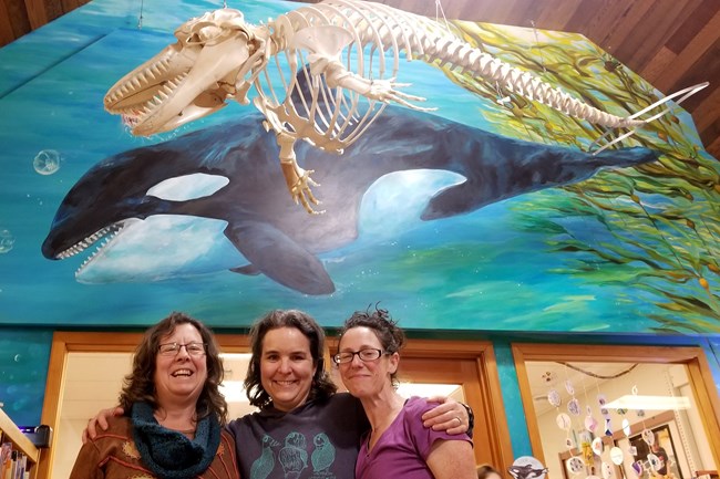 Mural artist and two NPS biologists pose under a mural and killer whale skeleton.