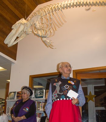 Two Tlingit tribal members stand wearing regalia under the newly installed killer whale skeleton