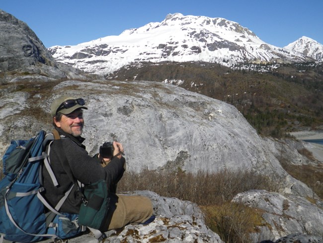 A man wearing a ball cap and sunglasses points his camera towards a rocky snow-covered mountain. He smiles at the camera.
