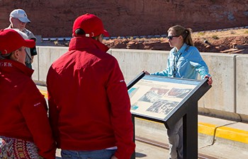 Tour guide stands behind interpretive sign while speaking to group atop dam