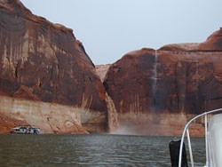 The bow of a boat looks at a muddy temporary waterfall off the side of a cliff into Lake Powell.