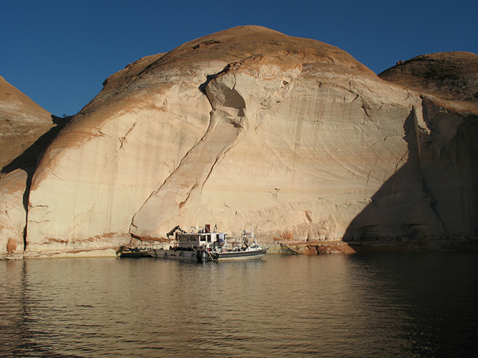 A large white boat sits in the middle of still water in front of a high canyon wall.