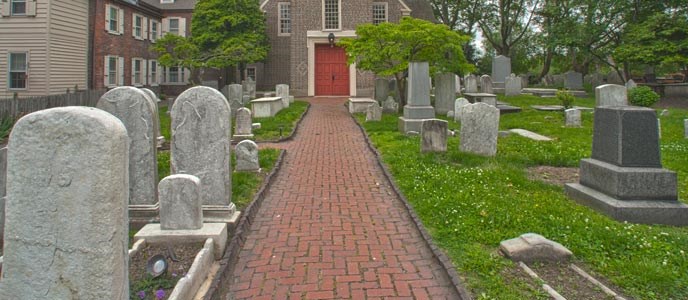 Color photo of a portion of the Gloria Dei cemetery with tombstones in the foreground lining both sides of a narrow brick walk that leads up to the red doors of the church in the background.