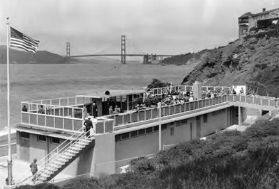 A beach house with visitors gathering on a viewing platform of an open roof balcony with the Golden Gate Bridge in the background.