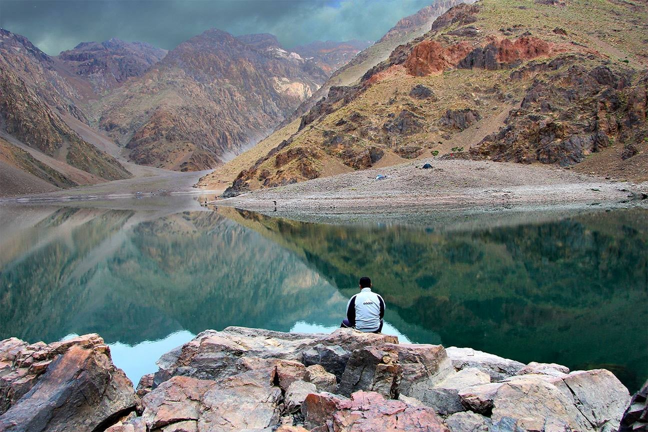 A visitor sitting in front of a serene alpine lake surrounded by mountains.
