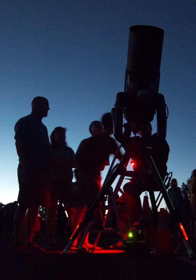 Red lights illuminate a large telescope. People gather around the telescope, trying to look through.