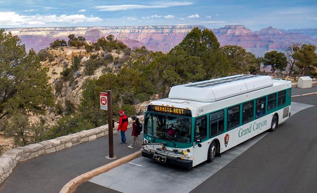 A white and green bus at a bus stop, In the background a paved walkway leads to a scenic overlook on the edge of a vast and colorful  canyon landscape.