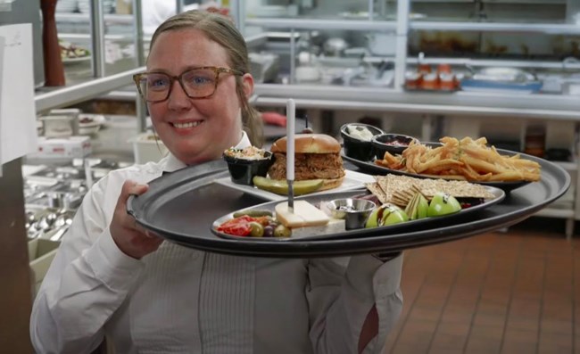 A woman food server is carrying a large tray of food out of a lodge kitchen