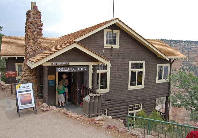 Dark brown house with white trim built into the edge of a cliff, with Grand Canyon in the background. A sign above the door reads "Kolb Studio"