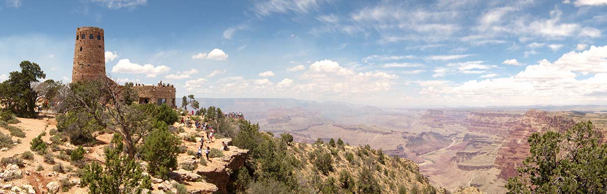 Expansive landscape of Grand Canyon on a hazy summer day. On the far left, the stone watchtower with many tourists walking around the canyon rim.