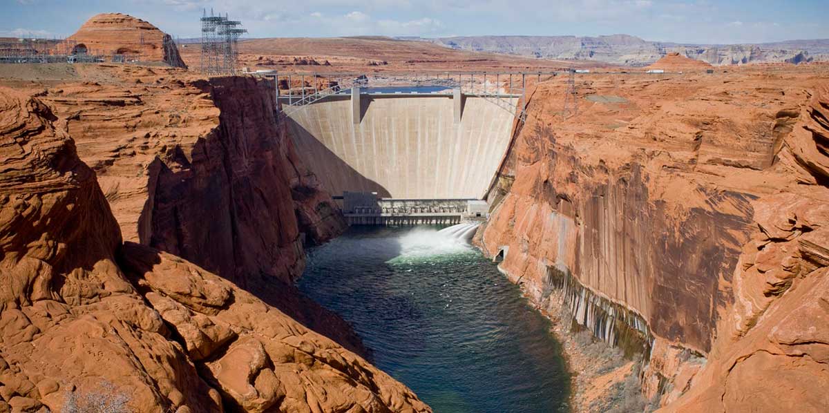 View upriver past beige colored sandstone cliffs on either side of Glen Canyon Dam. The water tubes at the bottom of the white , wedge-shaped dam are open and are discharging water jets for a high-flow event into the green-colored Colorado River.