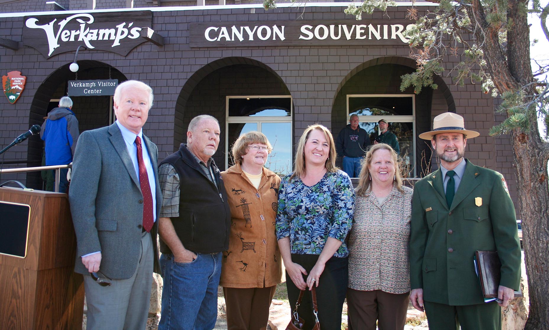 6 people posing for a group photo. 5 are members of the Verkamp family. On the far right is a man wearing a National Park Service uniform. The building in the background has signs that read, "Verkamp's", and "Canyon Souvenirs.