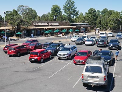 Looking over 2 dozen cars in a parking lot at a row of outdoor tables shaded by green umbrellas in front of a long building with lettering that reads, General Store.