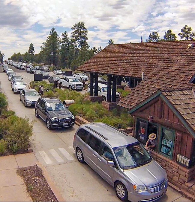 converging in the distance, three long lines of cars are waiting to purchase passes at a rustic entrance station. A ranger in the foreground is handing a map through a booth window to a family in a gray van.