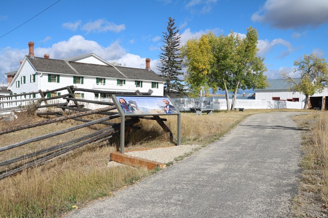 Wayside exhibit with level gravel landing off of smooth asphalt path.  The historic white ranch house is in the background.