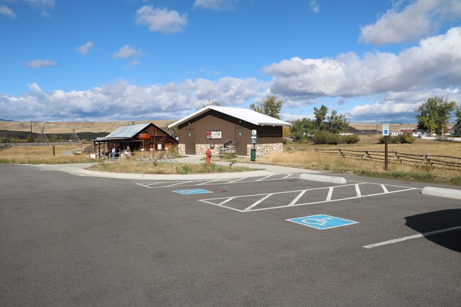 Asphalt path with 2 accessible parking spaces, building on front right with a door on each side to access restrooms.  Back left is visitor center building with a cement path connecting all things.