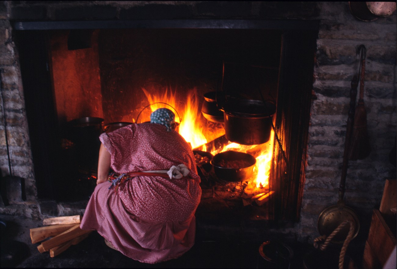 Fire in a stone hearth with a cook in period clothing leaning in.