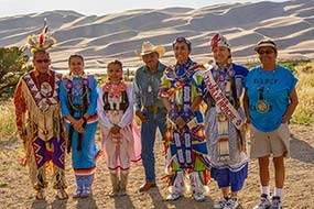 A group of Jicarilla Apache youth in colorful regalia with the dunes in the background