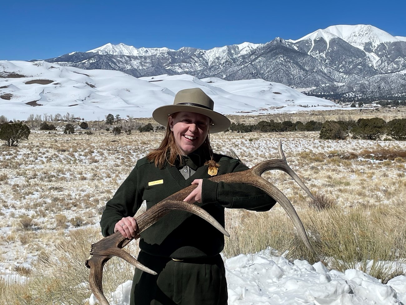 Ranger Grace holding an elk antler with snow covered mountains and sand dunes in the background