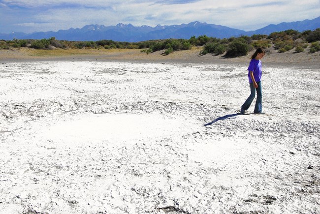 A girl walks across a deposit of white alkali, with dunes and mountains in the background