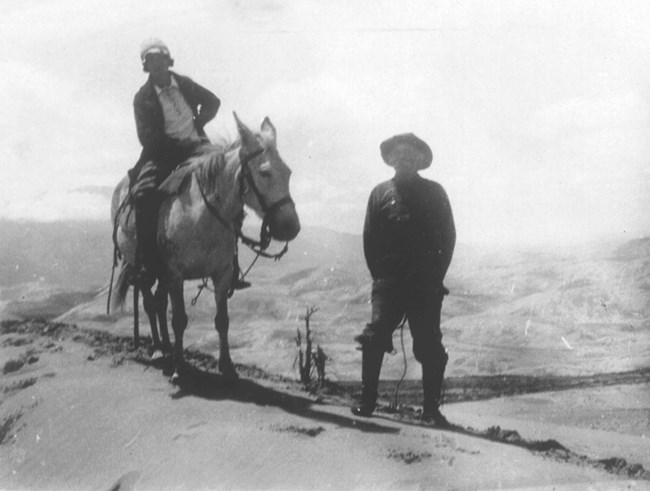 Historical black and white photo of a young woman on horseback with her father standing. They are on top of a dune with more dunes in the background.