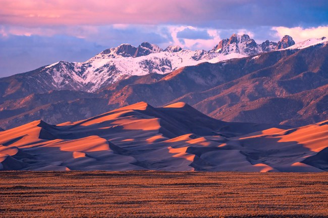 In sunset light are grasslands, a ridge of tall dunes, and jagged, snow-covered peaks.
