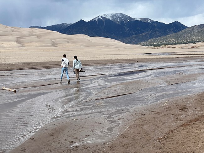 A narrow shallow stream flowing at the base of dunes and mountain with two people walking across it