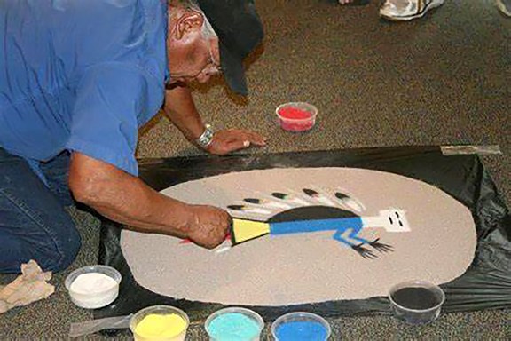 A Navajo (Dineh) man, Mitchell Silas, kneels over a sand painting of a stylized figure of blue, yellow, black and white, with feathers