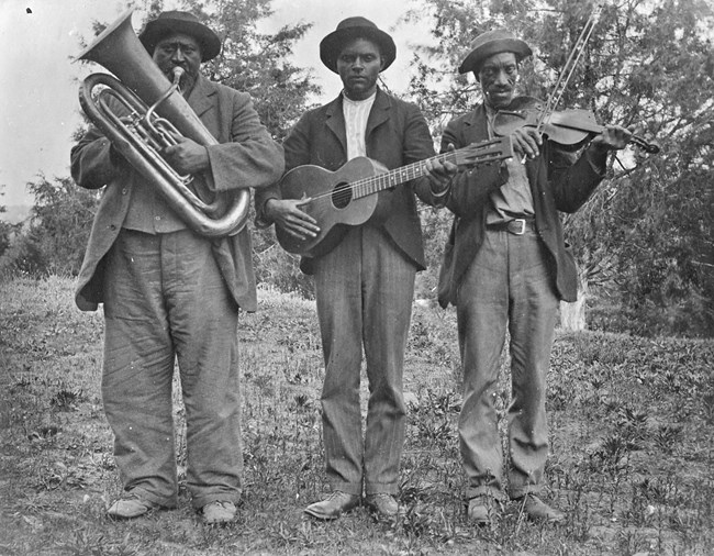 Three African American men are standing holding instruments - a tuba, guitar, and fiddle with a bow. They are each wearing a shirt, pants, coat, and a hat. They are standing outside with trees in the background.
