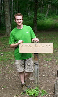 A backcountry hiker poses by a wooden directional sign located near a backcountry shelter.