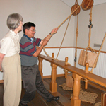 A volunteer looks on as a visitor uses a rope and pulley to lift a block of wood at the Naval Live Oaks Visitor Center.