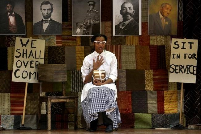 The image is of a play about African American history in the Midwest. The female actor is wearing period clothing of the 1950s. She is sitting on a bench with school books.