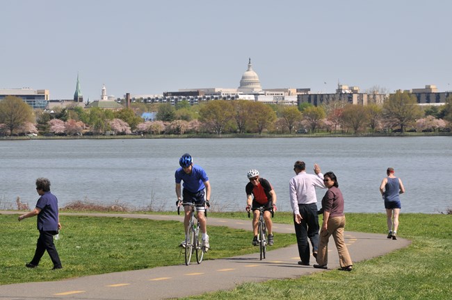 Pedestrians walk and cyclists ride along the Mt. Vernon Trail along the Potomac River with the United States Capitol in the background.