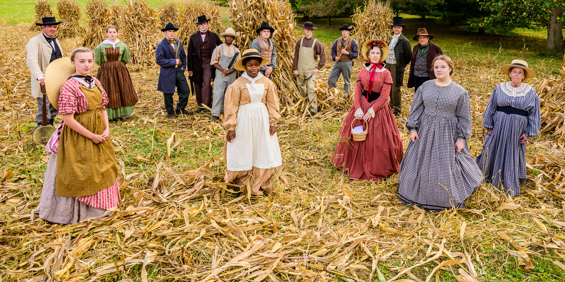 A group of living historians in 19th century clothing stand in a field of harvested corn.