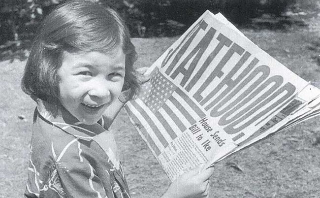 black and white image of a young girl reading a newspaper with the headline "Statehood"