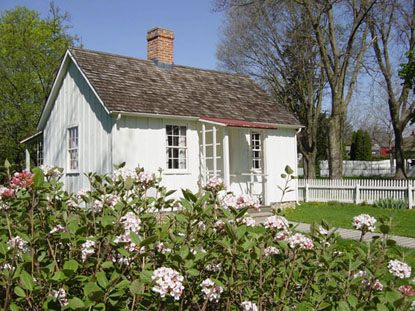 A small, white, frame cottage in the spring with a pink-flowering shrub in the foreground.