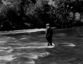 Black and white photograph of a man in a suit and hat fishing knee-deep in a stream.