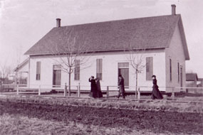 Black-and-white photograph of two couples in 19th century clothes approaching a white one-story building.