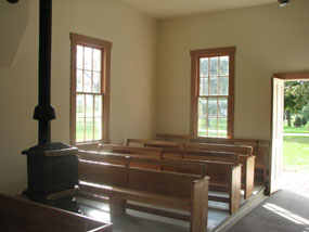Wooden benches and an iron stove in a sparsely furnished room are sunlit by double hung windows and an open doorway.