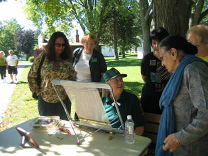 Karen Page Crislip demonstrates weaving on a small loom in the shade of a tree to six park visitors.