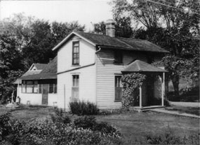 Black and white photo of the unrestored Birthplace Cottage: a two story white frame house.