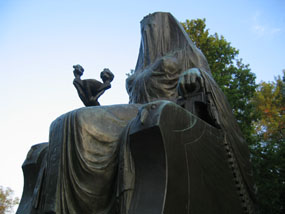 Bronze statue of a seated, veiled Egyptian goddess against the blue sky and trees of fall foliage.