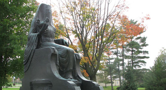 Bronze statue of a seated Egyptian goddess on a marble block with autumn foliage in the background.