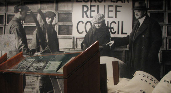A low-profile exhibit panel and stacks of flour sacks in front of a wall-sized black-and-white photo of Herbert Hoover with relief workers in Belgium.